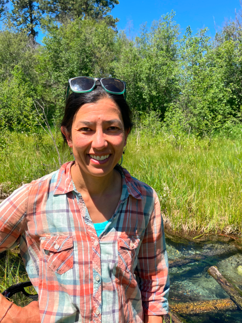 A woman wearing a pastel colored flannel shirt smiles and stands and smiles in front of shrubs, grass, and water.