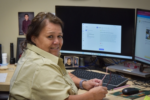 A woman with brown and hair wearing a tan, collared shirt sits in front of a computer and desk, looking at the camera and smiling.