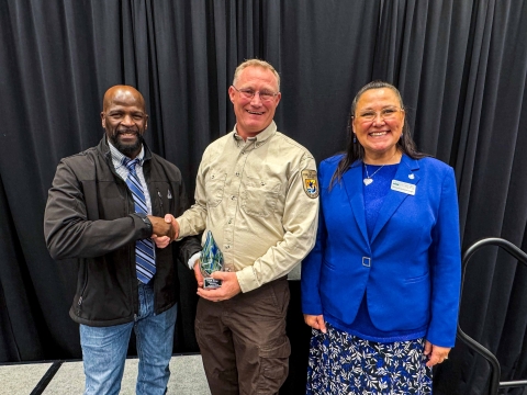 A unformed FWS employee shakes hands with another individual while holding an award. A woman stands smiling to the right of the awardee.