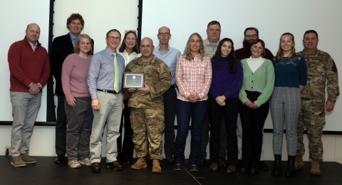 Croup of people, some in military camouflage uniforms, pose for a group photo. A man in a military camouflage uniform holds a plaque