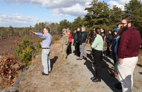 Group of people, some in military camouflage uniforms, outdoors looking off towards the left. A man in front has his arm extended as if he is showing something at a distance