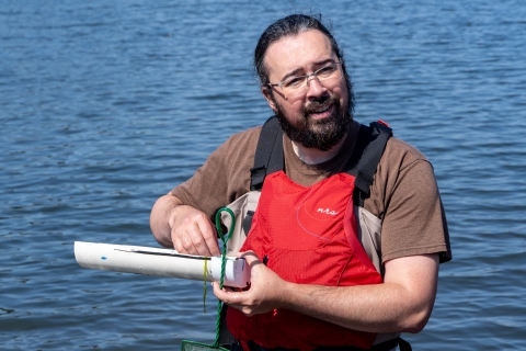 A service intern wearing waders and a PFD smiles as he measures the length of a fish. Water can be seen in the background.