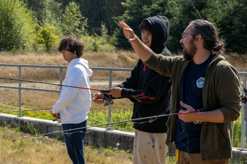 A service intern stands in a field with two teens; the three are all holding fishing rods. There is a railing and tree foliage in the background. 