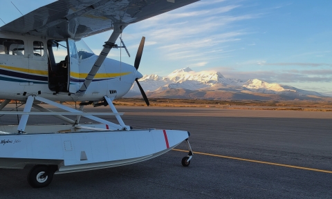 A small white airplane sits on a runway with snow capped mountains in the background