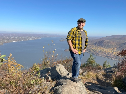 a white man wearing a brown newsboy cap, black and yellow checked shirt and jeans stands on a rock above a wide river with blue sky behind