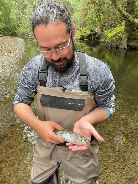 Service intern wearing waders and holding a cutthroat trout. A shallow river and tree foliage can be seen in the background. 