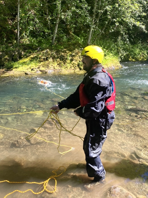 Service intern wearing a drysuit, PFD, and helmet and standing at a river edge pulling in the rope of a throw bag. 