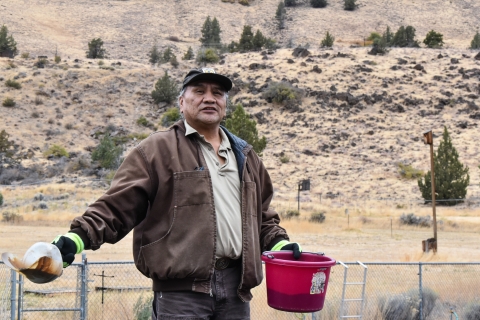 A man wearing brown and tan clothes holds a red bucket in one hand and a white scoop in the other.