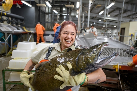 A woman with a huge smile on her face holds a massive salmon in both arms.