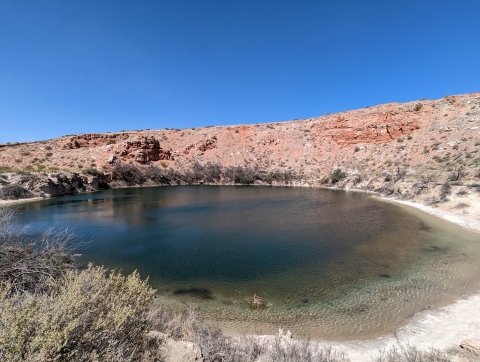 A shallow pool is encircled by rocky red bluffs and sagebrush on a bright sunny day.