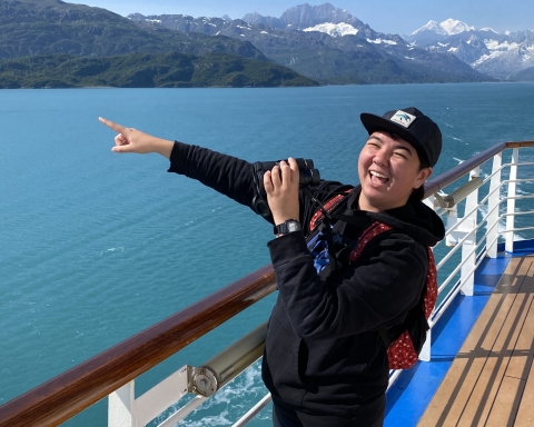 Ollie Zanzonico at Glacier Bay National Park. Ollie is standing on a boat pointing and smiling. There are snow-capped mountains in the background.