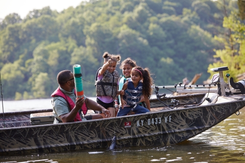 A man is fishing in a boat with three young girls. The kids are excitedly pulling a fish out of the water. 