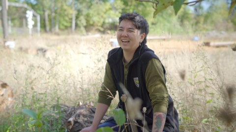 Ollie Zanzonico at Whitaker Ponds Nature Park in Portland, Oregon. Ollie is kneeing in the grass and smiling. They are wearing a black vest and olive green shirt.