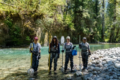 Four people stand at the shore of a river wearing waders and holding fishing nets as they smile at the camera. The shore is rocky, and trees can bee seen on the other side of the river.