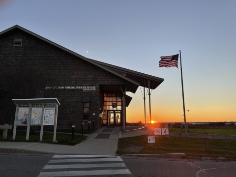 Sachuest Point visitor center, a brown building, with sun setting in background, and american flag on a flag pole blowing in the wind.