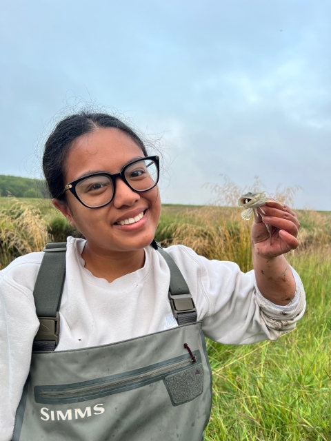 A Pathways employee smiles as she holds up a prickly sculpin. A field and sky can be seen in the background. 