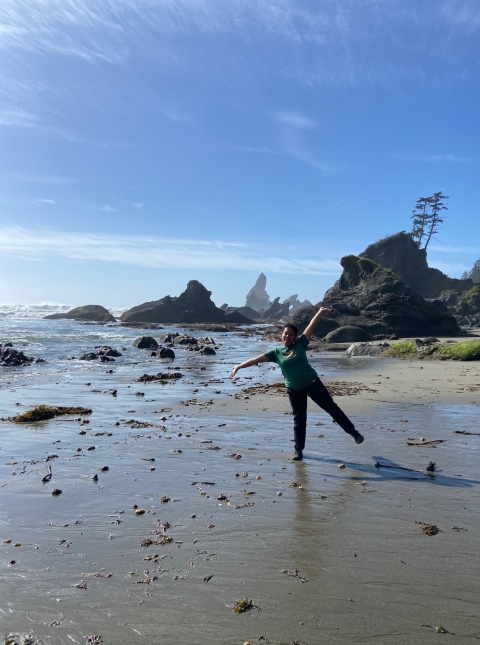 A Pathways employee smiles and strikes a pose on the beach. Large boulders are scattered in the background amongst the sand and water.