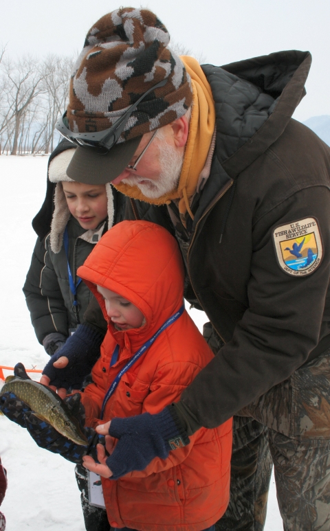 A US Fish and Wildlife Service staff member assists a child holding a fish, dressed in winter clothing