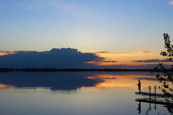 Silhouette of a person fishing from a pier against a sunset reflected on the water