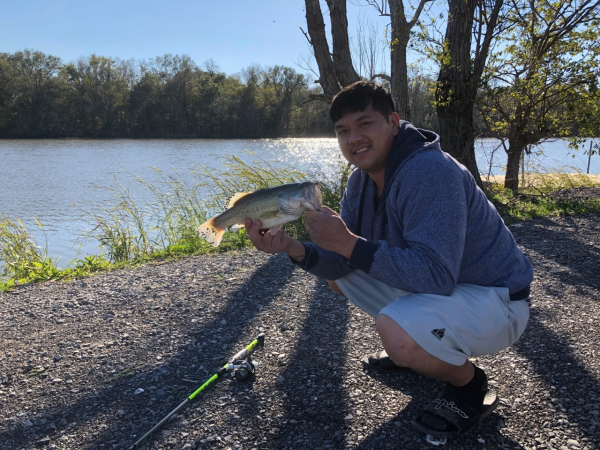 Man squatting next to a pond with a fishing pole holding a bass (fish)