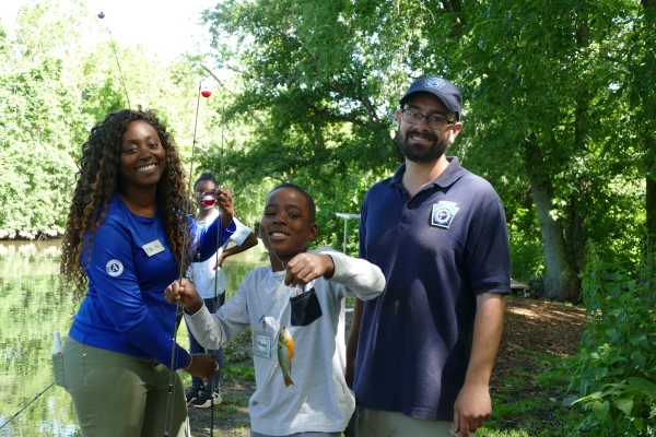 A smiling child holds a fish on a fishing line. Next to the child are two smiling refuge staff. 