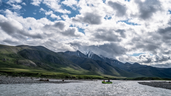 Two people float in a raft down a sparkling river surrounded by green mountains