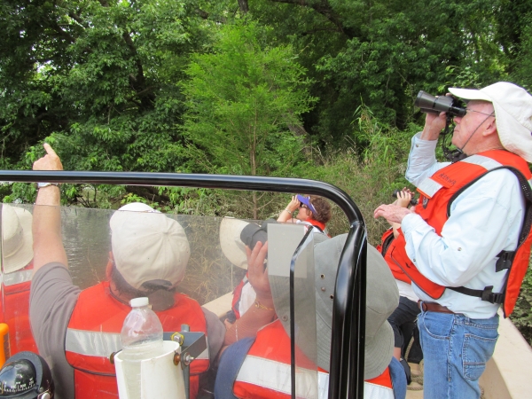 A group of birders wearing life vests in a boat point and look with binoculars at something in the trees. 