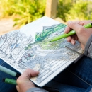 A hand holding a green felt tipped pen coloring in an illustration in a sketchbook of a mountainous scene