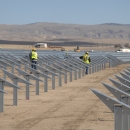 rows of gray solar panels stand out against the dry desert background. Solar panel installers wearing yellow vests work on the panels.