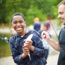 A boy holds a fish in his hands and smiles at the camera. A person next to him holds a fishing rod an bobber.