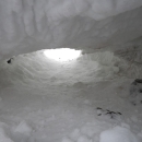 View from inside an empty polar bear den dug into the snow, looking out through the entrance hole toward the white sky.