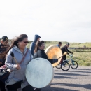 Woman with white skin drum leads group of people walking down a road. Kids on bikes flank her.