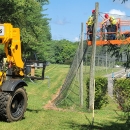 lush green trees surround a neatly trimmed grass field. A lowell (cherrypicker) forklift (10 ton heavy equipment) is on the left and a hemlock tree is on the right. A orange lift platform is suspended up from the right to the netting depicted in the middle along with the wood telephone poles that hold up the netting. There is a chain hanging on the lowell and two staff clad in yellow reflective jackets, red hardhats and dark blue pants on the lift platform about 15 feet in the air.