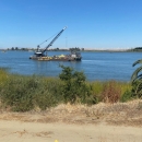 A large boat with heavy equipment in the water with greenery on the coast.