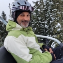 A man smiles while sitting on a ski lift wearing a helmet and green and white jacket. Trees with snow are in the background. 