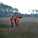 A man and a woman dressed in bright orange safety gear walk across a saltmarsh during a deer hunt
