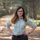 A woman with long brown hair stands on a trail framed by forest. Her hands are on her hips. She wears glasses and a U.S. Fish and Wildlife Service Uniform.