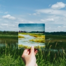 A hand holding up a tiny watercolor of the scene behind it