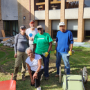 Five people stand in the yard of a building with wheelbarrows and other gardening tools