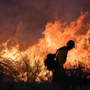 A firefighter is silhouetted in the dark walking along a fireline with orange flames in the background