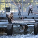 In the foreground, two staff at a fish hatchery work both above and in the water to remove some structures. Two people are in the background behind a chain link fence. 