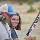 Youth participant in shooting sports holding pink firearm stands next to mentor. 