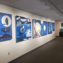 Photo of artwork hanging on walls in the Confluence Gallery that depicts various birds against water and starry skies.