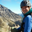 A woman wearing a backpack, rope, and climbing helmet sits high up with a mountain view in the distance