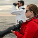 A woman wearing sunglasses and a red jacket standing in the foreground holding a camera with a large lens and looks out at sea on a ship. In the background, a man in a white jacket stands looking through a pair of binoculars.