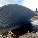 A huge culvert spanning a creek with a woman inside with outstretched arms and people walking on the road above