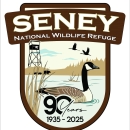 A shield logo showing the words Seney National Wildlife Refuge at the top. Centered in the logo is a Canada goose floating in the water with the words 90 years 1935 - 2025. In the background a fire tower stands, a black bear mother and two cubs walk along the edge of the pool, and two sandhill cranes in flight fly overhead.