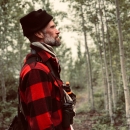 A side profile from the waist up, of a person wearing a red flannel shirt, a bear spray holster around their torso, and a black knit hat. They look away from the camera and off-frame with trees behind them..