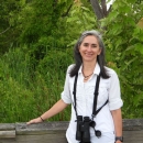 Person in white shirt stands at railing with greenery behind and binoculars hanging from neck. 