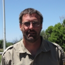 Man in a brown uniform with dark hair, glasses, and a beard
